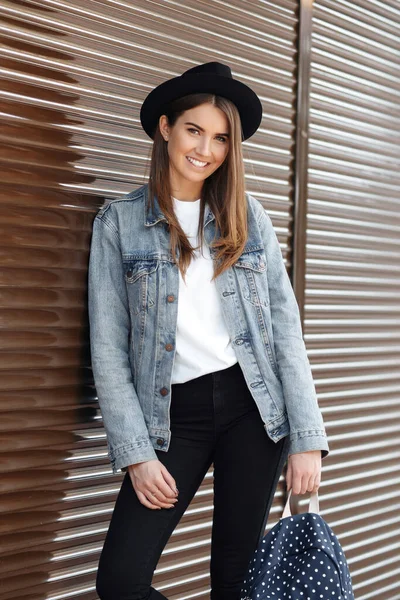 Glad leende hipster flicka med brunt hår bär hatt och ryggsäck utomhus. — Stockfoto