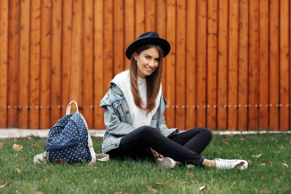 Hell lächelndes Hipstermädchen mit braunen Haaren mit Hut und Rucksack auf dem Rasen im Park sitzend. — Stockfoto