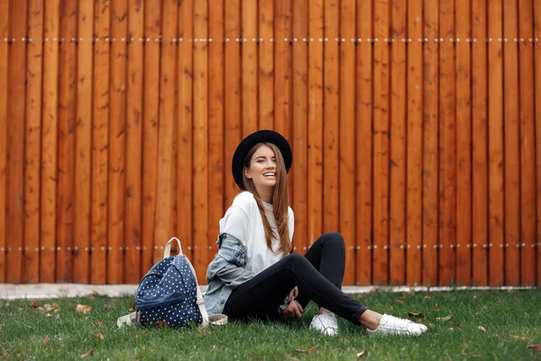 Elegante chica hipster con cabello castaño usando un sombrero y una mochila mientras está sentado en la hierba en el parque . — Foto de Stock