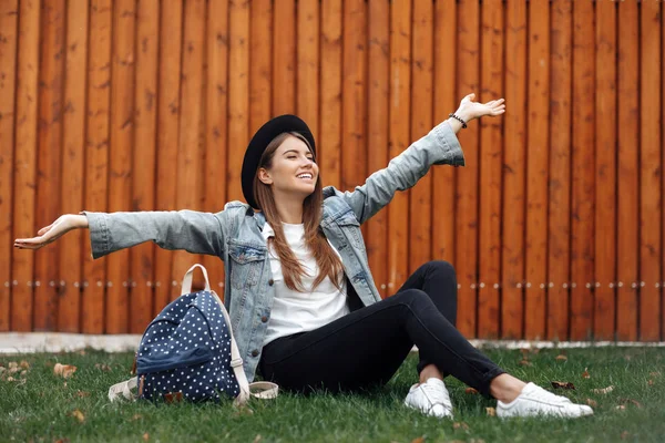 Chica hipster pacífica con un sombrero y una mochila mientras está sentado en la hierba con las manos abiertas en el parque . — Foto de Stock