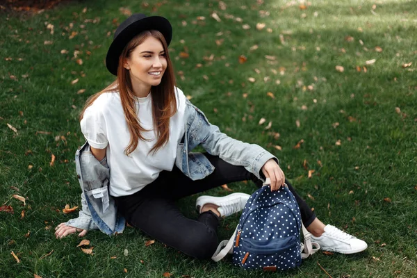 Hell lächelndes Hipstermädchen mit braunen Haaren mit Hut und Rucksack auf dem Rasen im Park sitzend. — Stockfoto