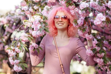 Charming young woman with curly pink hair and sunglasses near the blossoming spring tree.