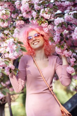 Bright woman with curly pink hair and sunglasses near the blossoming spring tree.