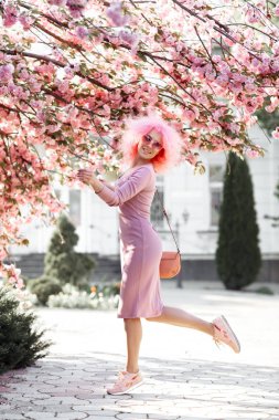 Full lengh photo of dancing young woman with curly pink hair and sunglasses near the blossoming spring tree.