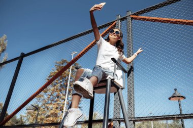 Hoş esmer, parlak gülüşlü, modern kıyafetler giymiş genç bir kadın stadyumda akıllı telefonuyla selfie çekiyor..