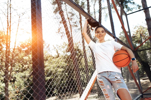 Yakışıklı, esmer, genç bir kadın. Parlak bir gülümsemesi var. Günlük modern kıyafetler giyip stadyumda basketbol oynuyor.. — Stok fotoğraf