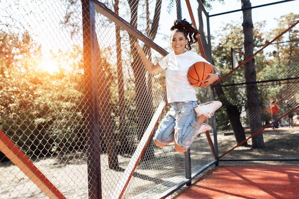 Oförsiktig brunett ung kvinna med ljusa leende klädd i casual moderna kläder hoppa med basket på arenan. — Stockfoto