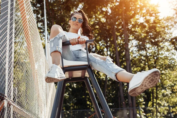 Atractiva joven con sonrisa brillante vestida con ropa moderna casual posando en silla de árbitro en el estadio . — Foto de Stock