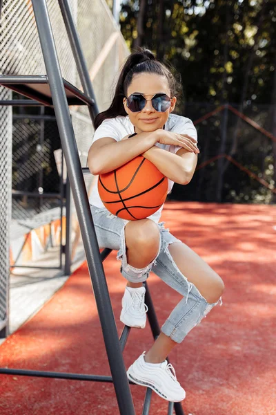 Modern kıyafetler giymiş şık genç bir kadın stadyumdaki hakem sandalyesinde basketbol topuyla poz veriyor.. — Stok fotoğraf