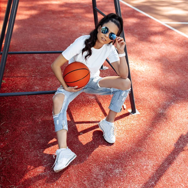 Esmer, modern kıyafetler giymiş, stadyumdaki hakem koltuğunda basketbol topuyla poz veren genç bir kadın.. — Stok fotoğraf
