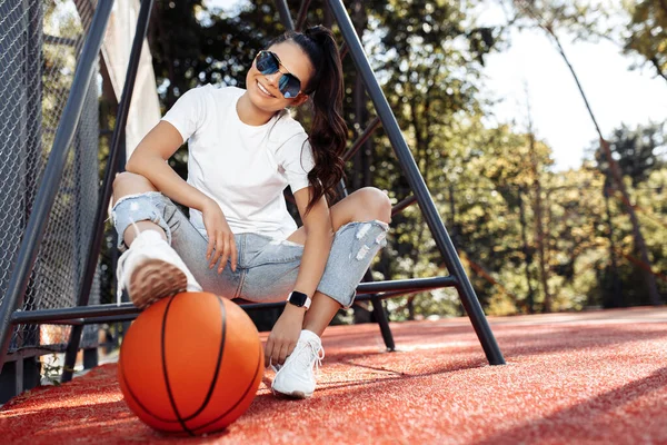 Brunett glad ung kvinna klädd i casual moderna kläder poserar med basket på arenan. — Stockfoto