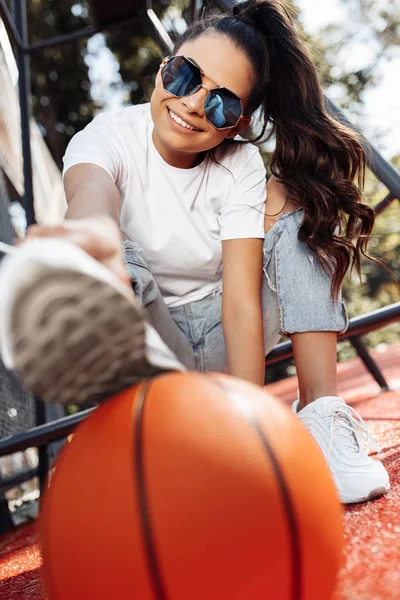 Gelukkig brunette jonge vrouw gekleed in casual moderne kleding en zonnebril poseren met basketbal in het stadion. — Stockfoto