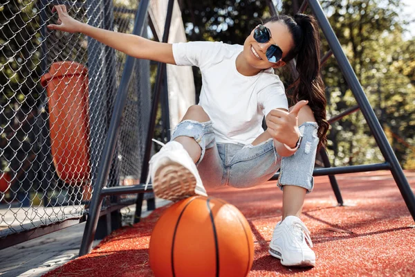 Brunett ung kvinna klädd i avslappnade moderna kläder poserar med basket på arenan. Ok gest. — Stockfoto