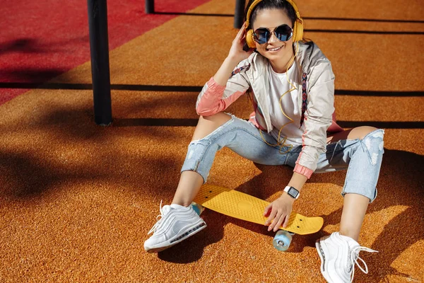 Glada brunett ung kvinna sitter och lyssnar musik som håller gul skateboard eller pennyboard på arenan. — Stockfoto