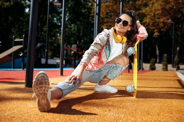 Trendig brunett ung kvinna sitter och lyssnar musik som håller gul skateboard eller pennyboard på arenan. — Stockfoto