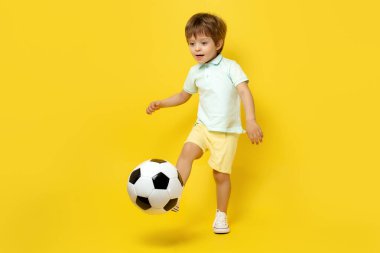 Yaz kıyafeti giymiş şirin bir çocuk sarı arka planda futbol topuyla oynuyor..