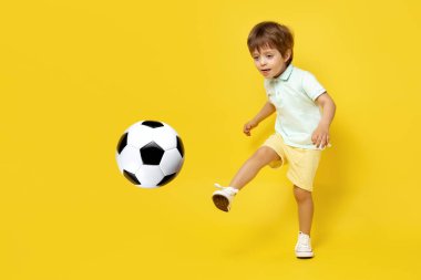 Sarı arka planda futbol topuyla oynayan sıradan yaz kıyafetleri içinde sevimli küçük bir çocuk..