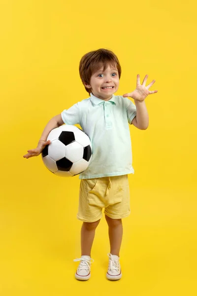 Αξιολάτρευτο μικρό παιδί σε casual καλοκαιρινό ντύσιμο κρατώντας μια μπάλα ποδοσφαίρου και ποζάροντας με αστείο πρόσωπο πάνω από κίτρινο φόντο. — Φωτογραφία Αρχείου