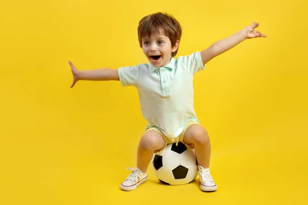 穿着休闲装的小男孩坐在足球上，胳膊伸到黄色的背景上，很可爱. — 图库照片