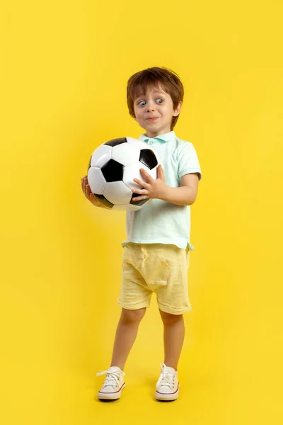 迷人的小男孩穿着休闲装，拿着足球，在黄色背景下摆出滑稽的姿势. — 图库照片