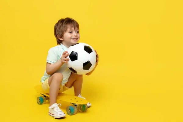 惊喜交集的小男孩坐在滑板上或黄底滑板上玩足球. — 图库照片