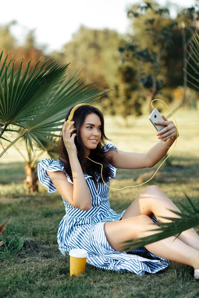 Jovem elegante no vestido de verão ouvindo música com fones de ouvido e tirar selfie no smartphone ao ar livre . — Fotografia de Stock