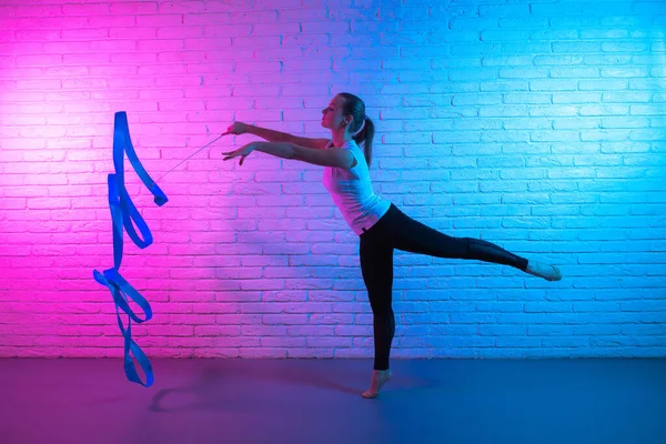 Turnerin, die sich vor einer Ziegelwand im Neonlicht dehnt. Flexible muskulöse Frau macht Spiralen mit Schleife. — Stockfoto