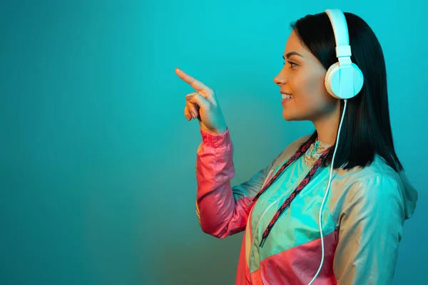 Schöne junge Frau im Sport-Outfit im 90er-Jahre-Stil mit weißen Kopfhörern, die im Studio im Neonlicht mit dem Finger zeigen. — Stockfoto