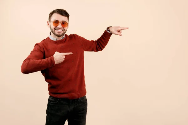 Vrolijk baard man in casual outfit dragen zonnebril wijzend vingers weg op beige achtergrond. — Stockfoto