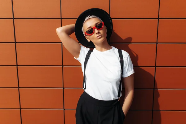 Sebedůvěra trendy hipster mladá žena na sobě černé sluneční brýle a klobouk skákání poblíž zdi moderní budovy. — Stock fotografie