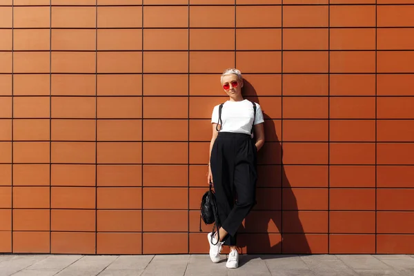 Mujer joven hipster serio con gafas de sol rojas y sombrero negro permanecer cerca de la pared del edificio moderno . — Foto de Stock