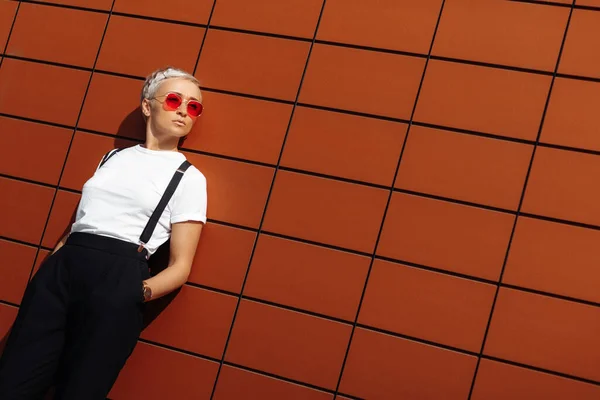 Krásná hipster mladá žena nosí červené sluneční brýle pobyt v blízkosti zdi moderní budovy. — Stock fotografie