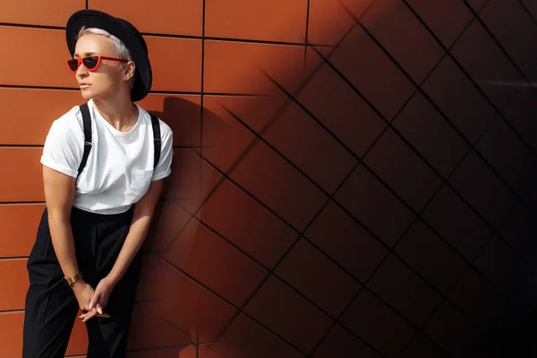 Joven hipster de moda con gafas de sol negras y sombrero saltando cerca de la pared del edificio moderno . — Foto de Stock