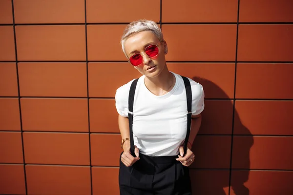 Stylový hipster mladá žena nosí červené sluneční brýle pobyt v blízkosti zdi moderní budovy. — Stock fotografie