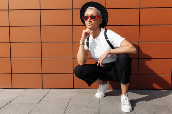 Hipster mujer joven con gafas de sol rojas y sombrero negro sentado cerca de la pared del edificio moderno . — Foto de Stock
