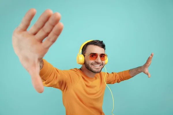 Knappe bebaarde jongeman met een zonnebril die naar muziek luistert met een gele hoofdtelefoon op een cyaanse achtergrond. — Stockfoto