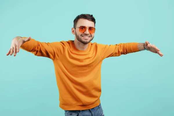 Knappe bebaarde jongeman met een zonnebril met wijd open handen op een cyaanse achtergrond. — Stockfoto