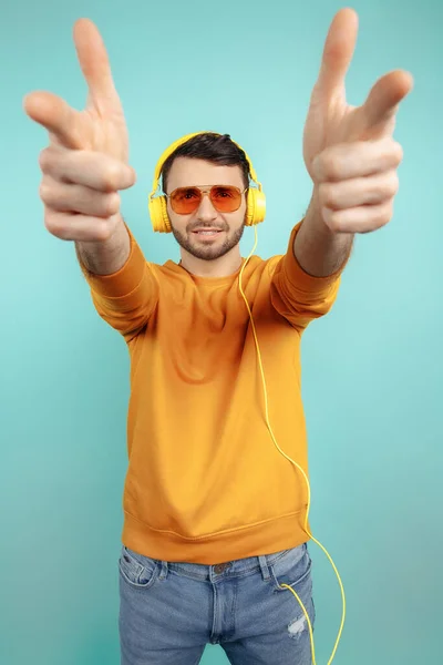 Zelfverzekerde bebaarde jongeman met een zonnebril die naar muziek luistert met een gele hoofdtelefoon op een cyaan achtergrond. — Stockfoto