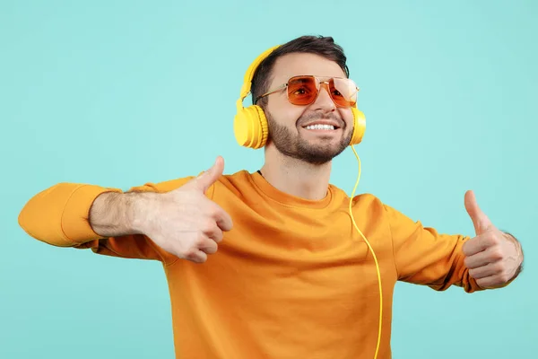 Vrolijke bebaarde jongeman met een zonnebril die naar muziek luistert die een goed gebaar op een cynische achtergrond laat zien. — Stockfoto