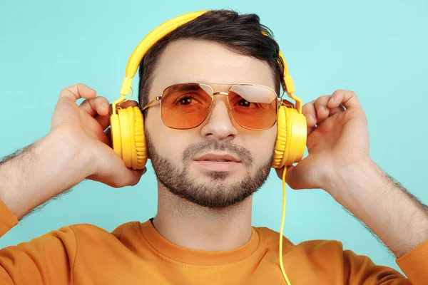 Verbazingwekkende bebaarde jongeman met een zonnebril die naar muziek luistert met een gele hoofdtelefoon op een cyaanse achtergrond. — Stockfoto