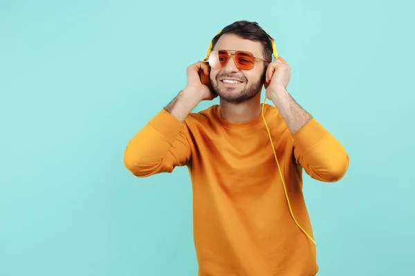 Verbazingwekkende bebaarde jongeman met een zonnebril die naar muziek luistert met een gele hoofdtelefoon op een cyaanse achtergrond. — Stockfoto