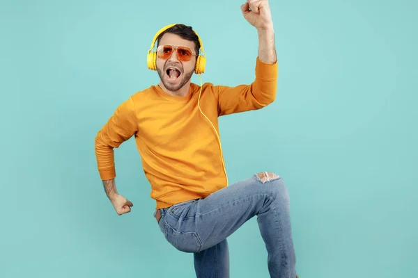 Emocionado joven barbudo con gafas de sol escuchando música celebrar el éxito sobre fondo cyan . — Foto de Stock