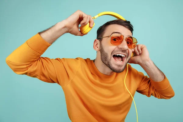 Mode bebaarde jongeman met een zonnebril die naar muziek luistert met een gele hoofdtelefoon op een cyaanse achtergrond. — Stockfoto