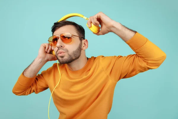 Förbluffad skäggig ung man bär solglasögon lyssna på musik med gula hörlurar på cyan bakgrund. — Stockfoto