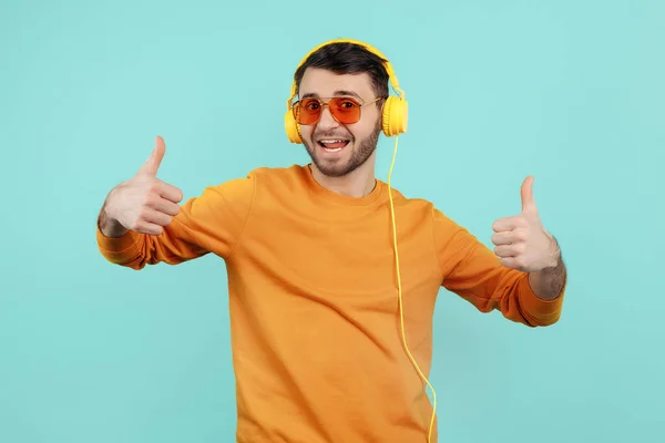 Gelukkig bebaarde jongeman draagt een zonnebril luisteren naar muziek tonen ok gebaar op cyaan achtergrond. — Stockfoto