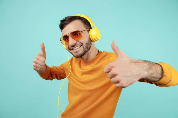 Blij bebaarde jongeman draagt een zonnebril luisteren naar muziek tonen ok gebaar op cyaan achtergrond. — Stockfoto