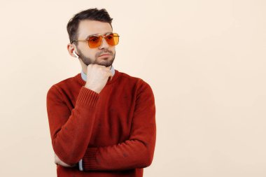 Ciddi sakallı, sarı güneş gözlüğü ve bej arka planda bluetooth beyaz kulaklık takan genç adam..