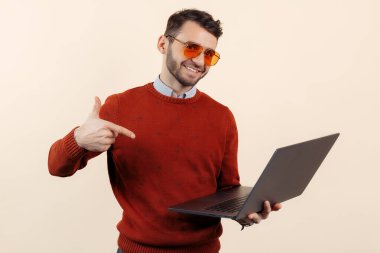 Sıradan kıyafetli şık sakallı genç adam bej arka planda bir bilgisayarı işaret ediyor..
