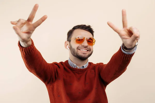 Vrolijke bebaarde man in casual outfit met een zonnebril met vredesgebaar op beige achtergrond. — Stockfoto