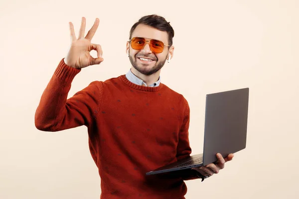 Glimlach bebaarde jongeman in casual outfit met notebook of computer show ok gebaar over beige achtergrond. — Stockfoto